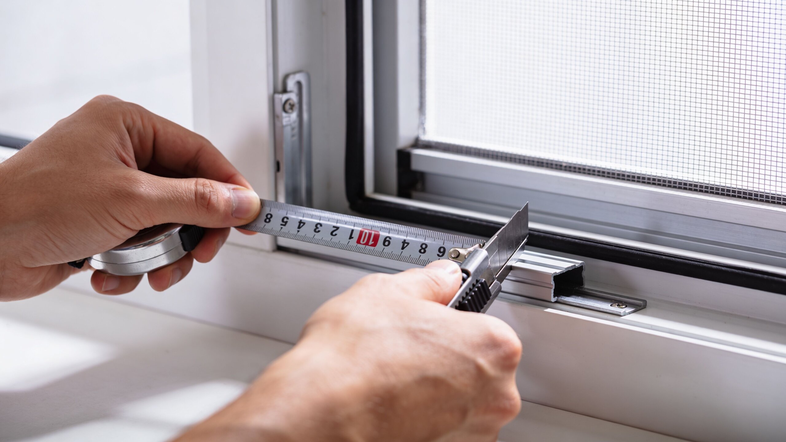 A person measuring a window frame with a tape measure to prepare for installing window screens.