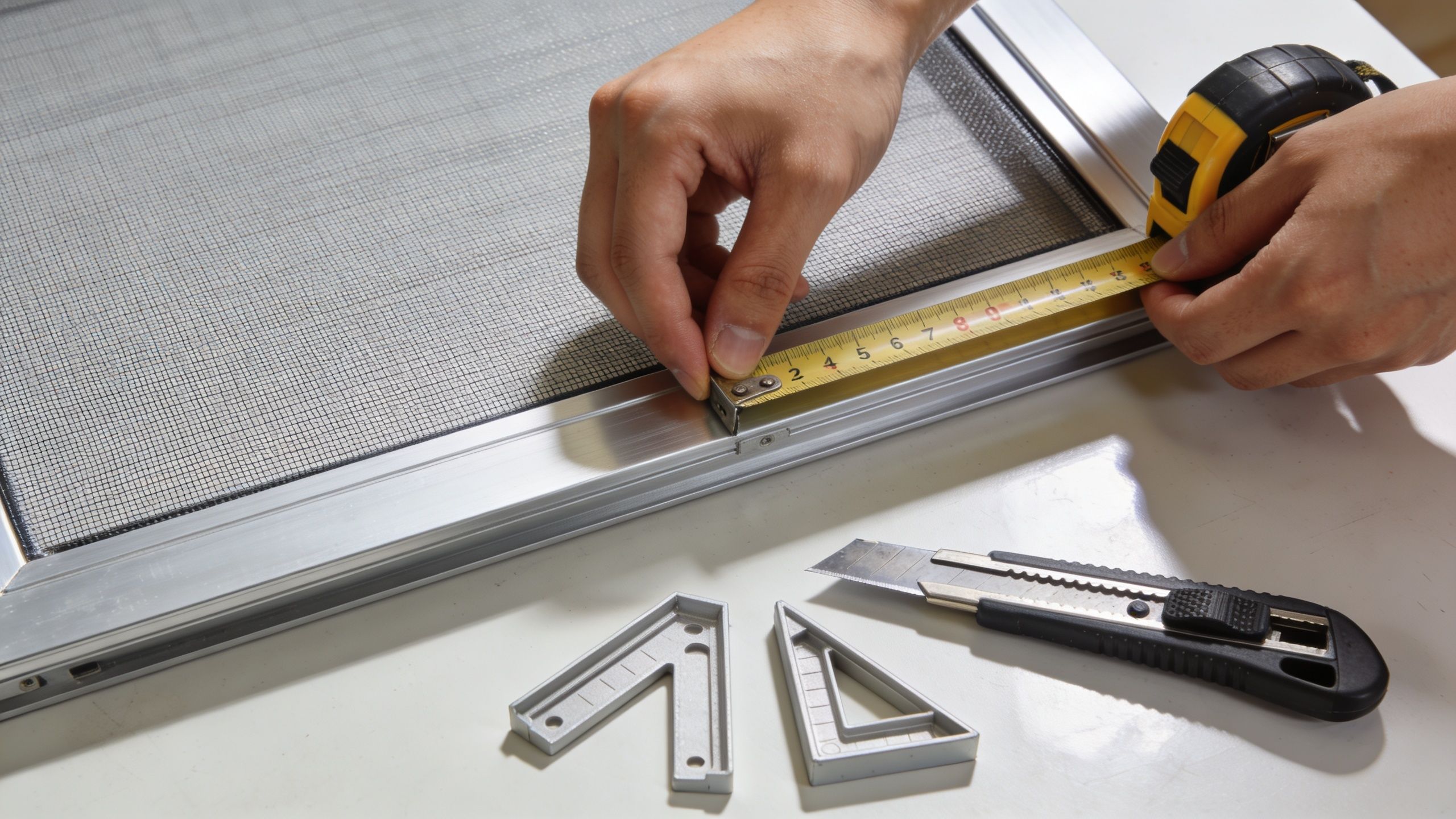 A person measures the width of a metal-framed window screen with a yellow measuring tape.