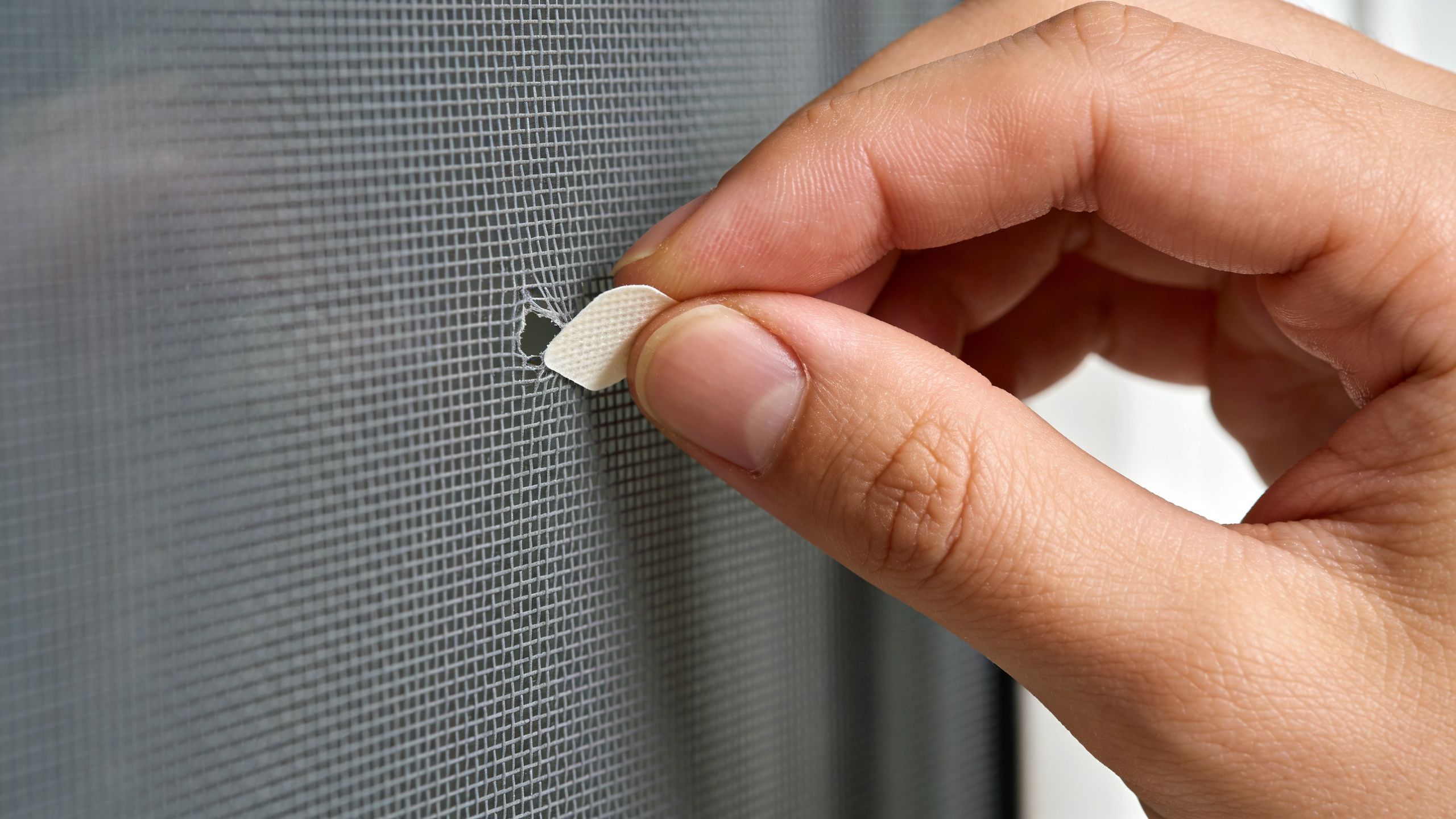A close-up view of a person using an adhesive patch to repair a hole in window screening.