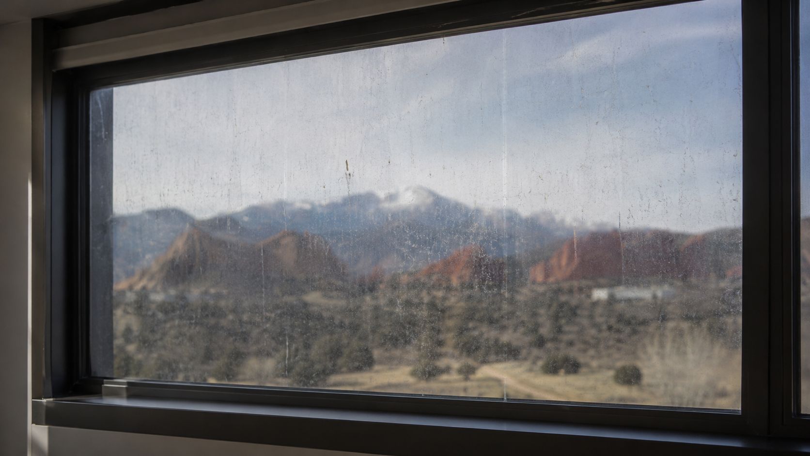 A view of snow-capped mountains and red rock formations seen through a dusty, streaky window pane.