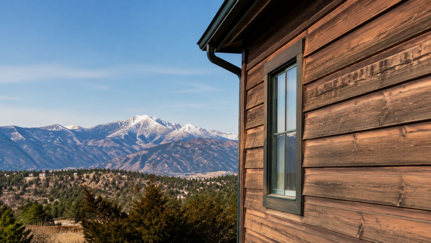 A rustic wooden cabin wall with siding, featuring a window overlooking a scenic mountain range view.