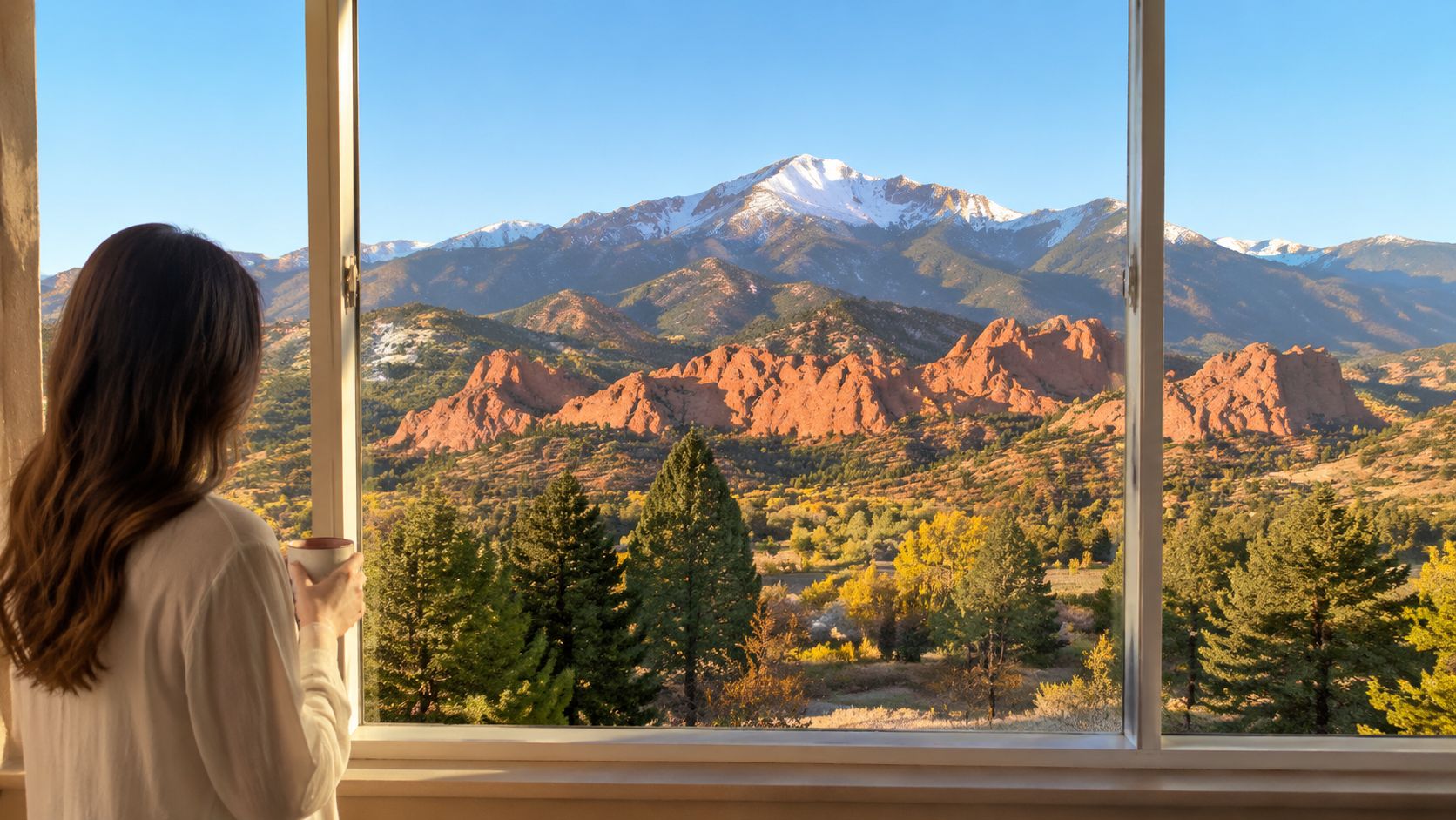 A woman holding a mug looks out a large window at a stunning mountain landscape with red rocks.